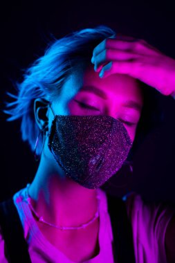 Gece kulübünde sahte elmaslarla dans eden koruyucu maskeli, güzel, modaya uygun genç bir kadın. Neon renkli ışık. Moda modelinin yakın plan portresi.