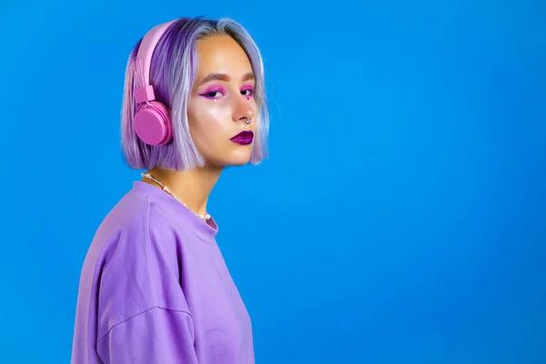 Hezká dívka s obarvenými fialovými vlasy, poslouchající hudbu, usmívající se, tančící ve sluchátkách ve studiu na modrém pozadí. Hudba, tanec, rozhlasový koncept — Stock fotografie