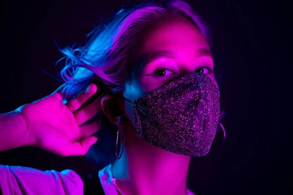 Gece kulübünde sahte elmaslarla dans eden koruyucu maskeli, güzel, modaya uygun genç bir kadın. Neon renkli ışık. Moda modelinin yakın plan portresi.