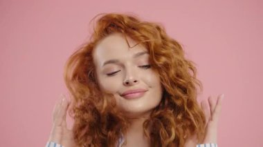 Oyuncu kıvırcık saçlı, gülümseyen, pembe stüdyoda kameraya kur yapan kadın. Çekici, modaya uygun, şık giyinen bayan uzun, harika saçlarını düzeltiyor. Yüksek kalite 4k görüntü