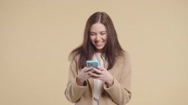 Akıllı telefondan internette sörf yapan güzel bir kadın. Bej arka planda gülücük ve neşe dolu bir kız. Teknoloji, başarı, mutluluk, sosyal ağ konsepti. Yüksek kalite 4k