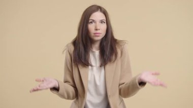 30 'lu yaşlardaki güzel iş kadını omuzlarını kaldırıyor. Yardımcı olamıyor. Bilmiyorum jestler yapıyor. Zor soru, bej arka planda suçlu tepkisi. Yüksek kalite 4k