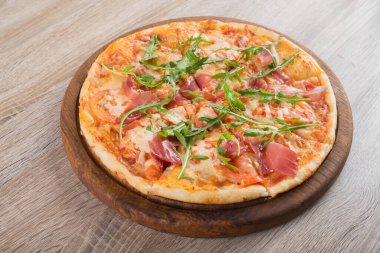 Tahta masa üstünde jambonlu, domatesli ve yeşil roka soslu taze pizza.