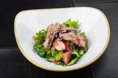 Beyaz tabakta dana eti ve susam tohumlu salata, siyah arka plan.