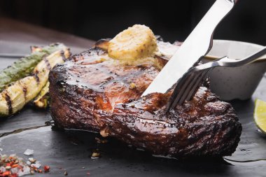 Baharatlı, çatallı ve bıçaklı ızgara biftek tişörtü. Siyah tahtada. Lezzetli restoran yemekleri