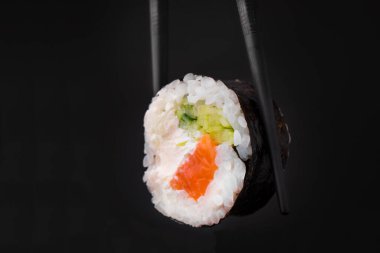 Somon ve salatalıklı bir suşi rulosunun yakın plan fotoğrafı siyah arka planda yemek çubuklarıyla tutulur.