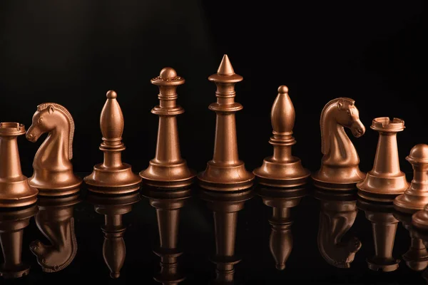 A series of gold chess pieces on a black glossy surface. Game set on dark background.