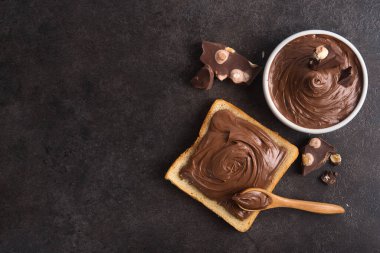 Çikolata, kaşıkla bir dilim ekmeğe, erimiş krema beyaz kâsesine ve taş bir masada fındıklı çikolataya sürüldü. Popüler tatlı yiyecekler. Üst görünüm.