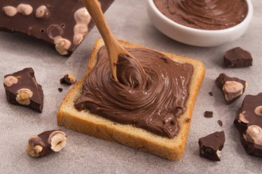 Çikolata, kaşıkla bir dilim ekmeğe, erimiş krema beyaz kâsesine ve taş bir masada fındıklı çikolataya sürüldü. Popüler tatlı yiyecekler.