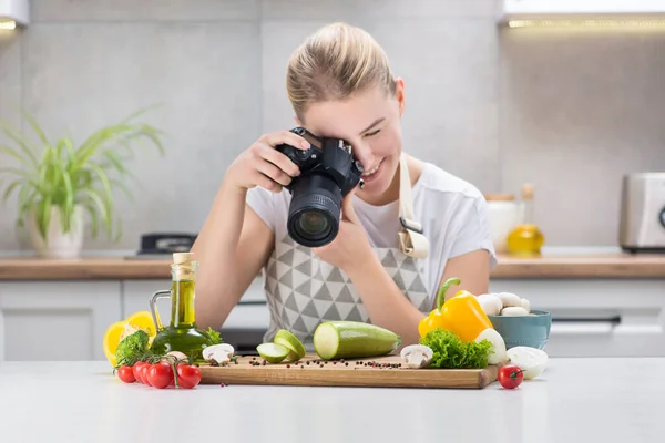 Female food blogger shoots content on the camera in kitchen with fresh vegetables on a table. Blog about healthy eating. Step by step recipes dishes.