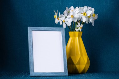 Sarı İskandinav tarzı vazoda güzel narsis çiçekleri ve ptoho için boş çerçeve, arka planda mavi kumaş. Aile köşesi.