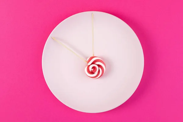 Time to start diet and give up sweet harmful, high-calorie foods. Plate with clock hand candy on pink background. Top view.