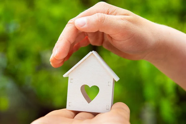 Small white model of house with heart window stand on child hand at green outdoors background.