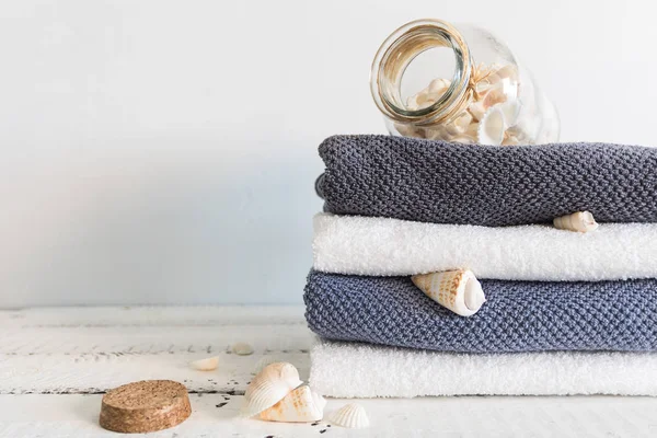 Uma pilha de toalhas de banho brancas com fita azul e diferentes conchas perto do frasco com conchas na mesa de madeira branca. Spa e bem-estar, algodão têxtil terry . — Fotografia de Stock