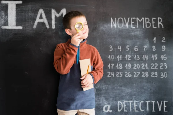 Jonge jongens permanent met Vergrootglas en notebook in zijn handen in de buurt van blackboard. Jonge detective. Creatief ontwerpconcept voor 2019 kalender. November — Stockfoto