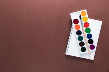 Üstten görünüm muhteşem bir boya, fırça ve defter üzerinde kahverengi bir arka plan üzerinde. Geri okul kavramı. Ofis malzemeleri düz yatıyordu.