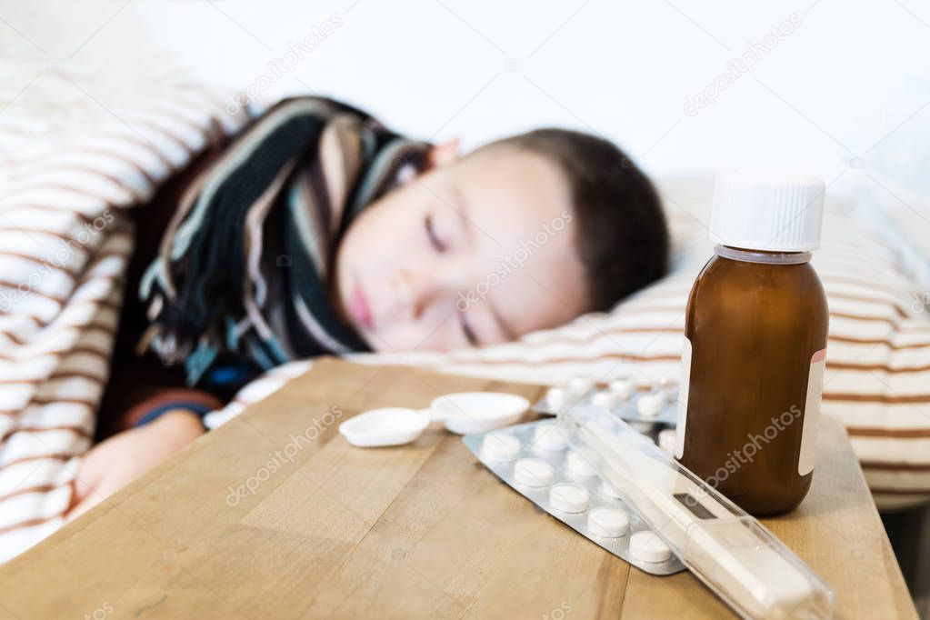 Young boy wearing sweater and a scarf sleeping in his bed while having a flu. Meds and syrips on a wooden table in front of him. Flu and seasonal diseases concept. Selective focus