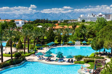 ANTALYA, TURKEY - 13 Nisan 2016: Yüzme havuzu, palmiye ağaçları ve çamları olan IC Hotels Green Palace 'ın bölgesi. Turistler şemsiyelerin altında güneşlenirler. Arka planda dağlar ve bulutlar.
