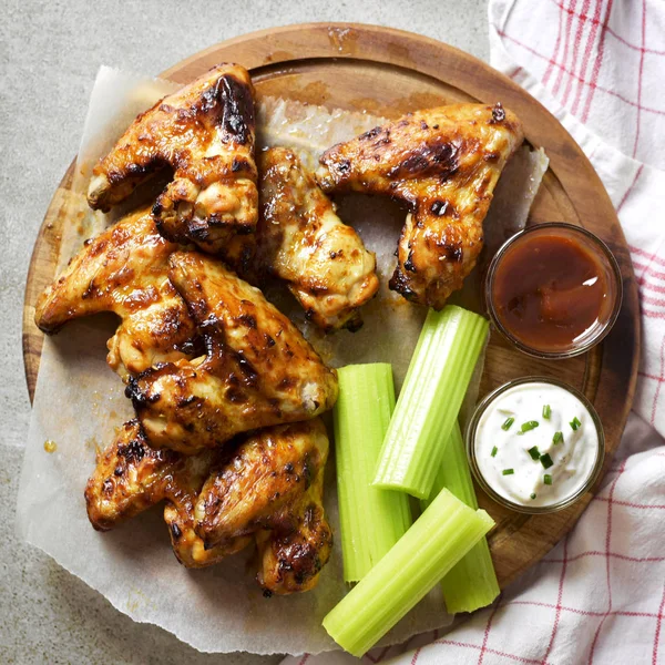 Delicious chicken wings on a wooden cutting board. Close-up shot of tasty chicken wings with perennial celery, sour cream dip and bbq sauce.