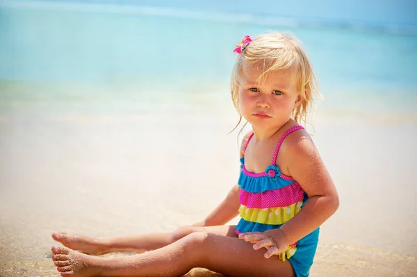 一个可爱的漂亮孩子在海滩上的肖像 可爱的小金发女孩穿着漂亮的彩色泳装坐在沙滩上 — 图库照片