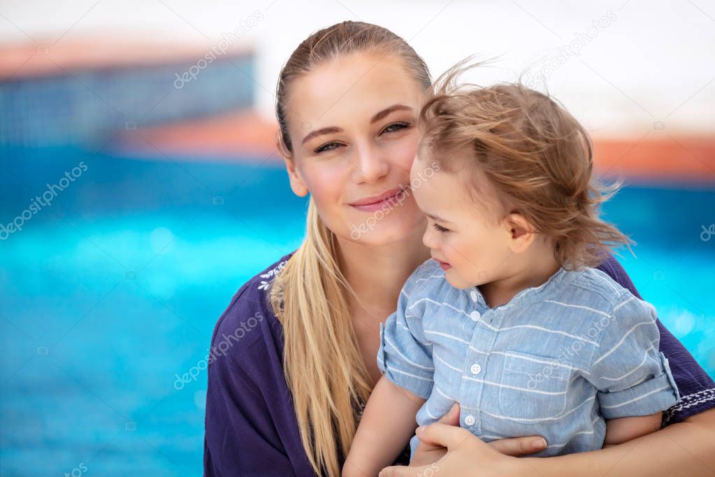 Portrait of charming mother with cute little son sitting near poolside and enjoying warm sunny day, spending summer holidays on the beach resort