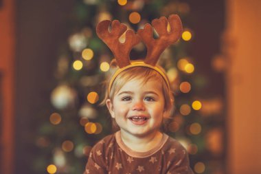 Rudolphs 'un ren geyiği kostümü giyen neşeli bir bebeğin portresi, Noel' i evde güzel, parıldayan bir Noel ağacının yanında geçirmek zevkle.