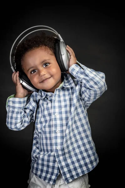 かわいいアフリカ系アメリカ人の少年が大きなイヤフォンを持っていて 喜びの音楽を聞いて スタジオの黒い背景の上に立っている肖像画 — ストック写真