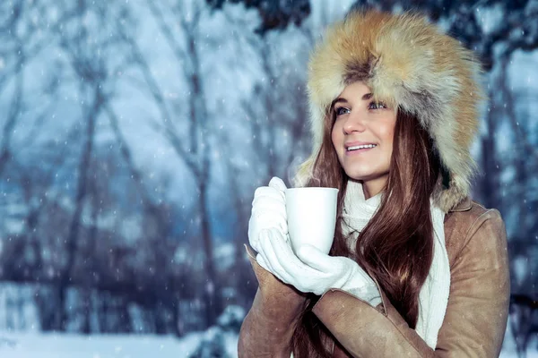 一个年轻貌美的姑娘 戴着一顶时髦的帽子 站在雪地的公园里 温热的手捧着热腾腾的茶杯 欣赏着美丽的冬季美景 — 图库照片