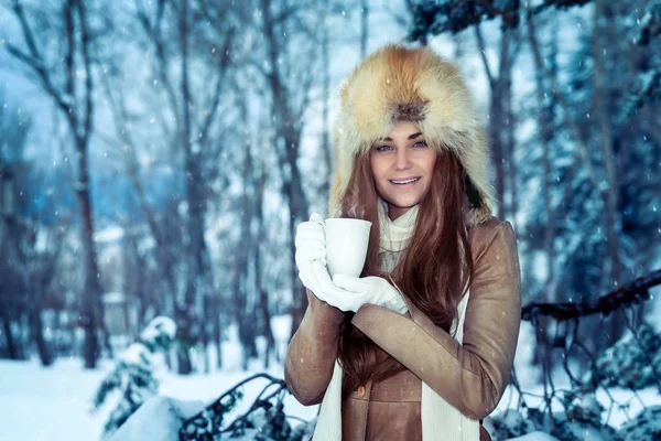 雪の公園に立ち 冬の休日を楽しむ手にお茶の熱いカップとスタイリッシュな毛皮の帽子を身に着けている美しい女性の肖像画 — ストック写真