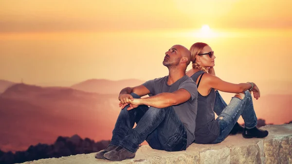 Happy couple enjoying sunset view, handsome guy with girlfriend sitting back to back to each other over mountains background in mild orange light, romantic vacatio