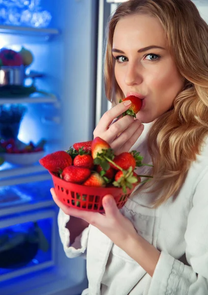 オープン冷蔵庫の近くに立っている美しい女性の肖像画とイチゴを食べます 喜びは新鮮な甘い果物をかむと 健康的な有機栄養 — ストック写真