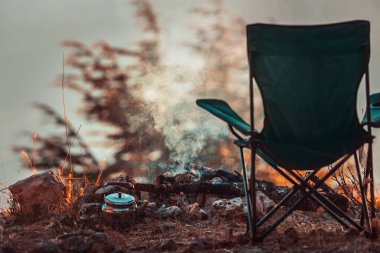 Bir Sonbahar Huzurlu Eğlencesinin Kavramsal Fotoğrafı. Kamp ateşinde Teapot 'un yanındaki Boş Kamp Sandalyesi. İnsan yok. Doğadaki Huzur ve Gevşeme.
