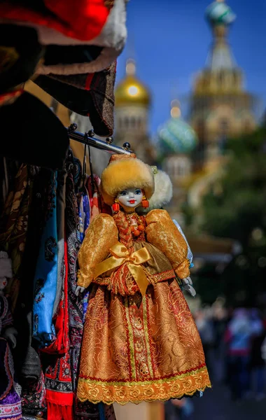 Russian doll dressed in a traditional winter outfit with a fur hat in on display at a souvenir shop in an open air market with the Church of Savior on Spilled Blood in the background in Saint Petersburg Russia