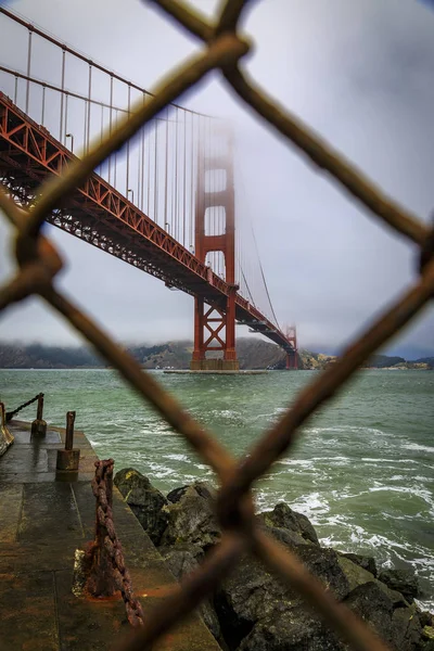 The famous Golden Gate bridge viewed through a rusty chain link fence on a cloudy day in San Francisco, California