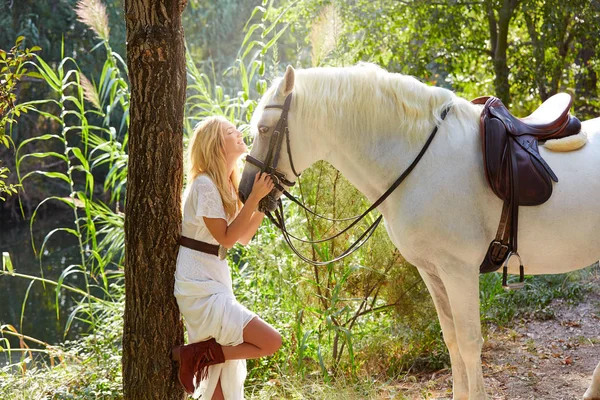 ブロンドの女の子が川の近く魔法のライトの森で彼女の白い馬を抱っこ — ストック写真