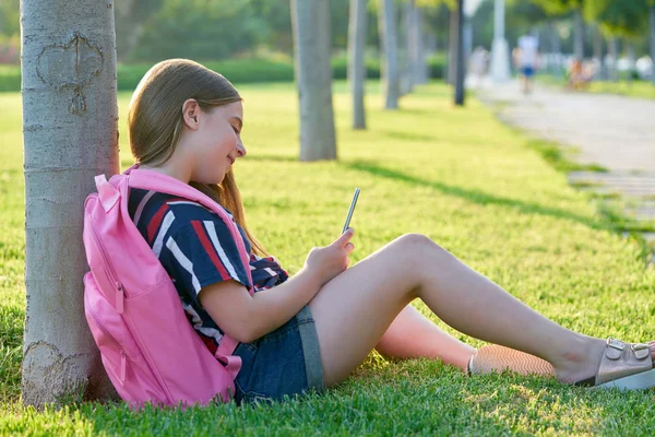 金发的学生女孩与智能手机在公园回学校坐在草地上 — 图库照片