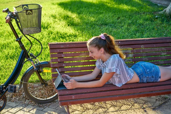 Mädchen Liegt Auf Parkbank Und Spielt Mit Tablet Und Fahrrad — Stockfoto