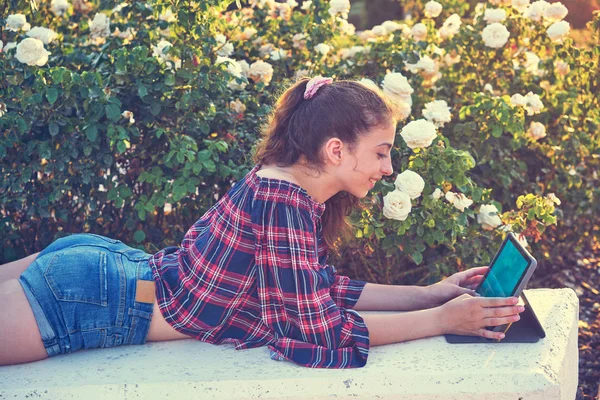 女孩躺在公园长椅上玩平板电脑在玫瑰花背景 — 图库照片