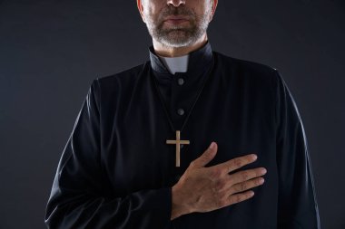Priest hand in heart gesture with cross on black background clipart