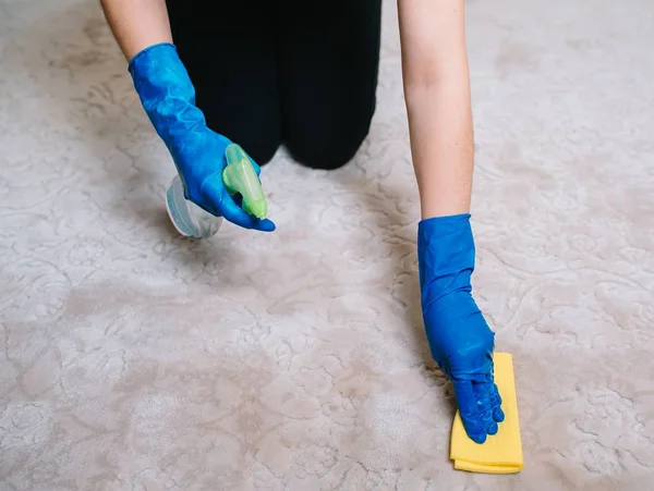 Las personas, las tareas domésticas y el concepto de limpieza: primer plano de la mujer en guantes de goma con tela y alfombra de limpieza en aerosol derergente en el hogar. primer plano de la mujer con alfombra de limpieza de tela — Foto de Stock