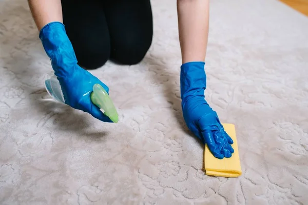 Las personas, las tareas domésticas y el concepto de limpieza: primer plano de la mujer en guantes de goma con tela y alfombra de limpieza en aerosol derergente en el hogar. primer plano de la mujer con alfombra de limpieza de tela — Foto de Stock