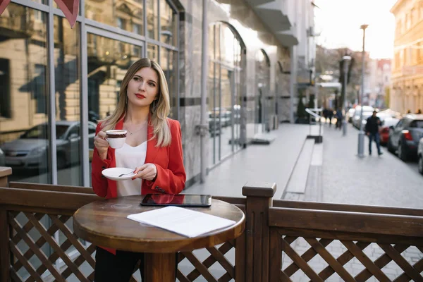 Hermosa joven empresaria con una taza de café desechable, bebiendo café y sosteniendo la tableta en sus manos contra el fondo urbano de la ciudad . — Foto de Stock