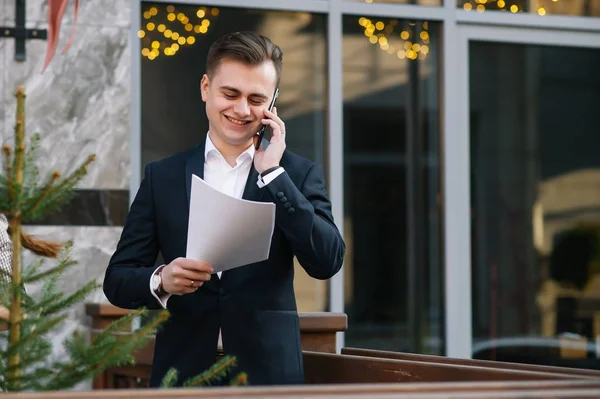 Jeune homme d'affaires avec documents et téléphone portable près du bâtiment moderne . — Photo