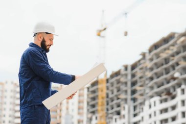 İnşaatçı inşaat halindeki eve bakıyor. Elinde çizimler olan Muchin şantiyeye bakıyor. Kameraya arkası dönük inşaatçı. Bina üniformalı ve koruyucu kasklı bir adam.