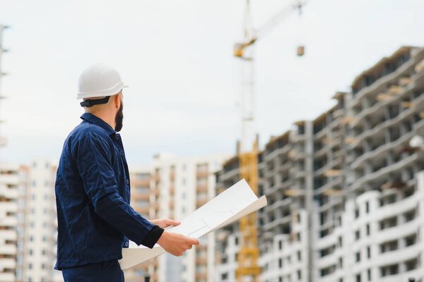 Builder looks at the house under construction. Muchin with drawings in his hands looks at the construction site. Builder with his back to the camera. Man in a building uniform and a protective helmet