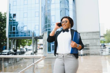 Beyaz tişörtlü başarılı Afro-Amerikan kadın çalışanın yarım boy portresi. Elinde akıllı telefon ve klasörle ofis binasının yanında kameraya gülümserken elinde belgeler var.