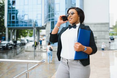 Beyaz tişörtlü başarılı Afro-Amerikan kadın çalışanın yarım boy portresi. Elinde akıllı telefon ve klasörle ofis binasının yanında kameraya gülümserken elinde belgeler var.