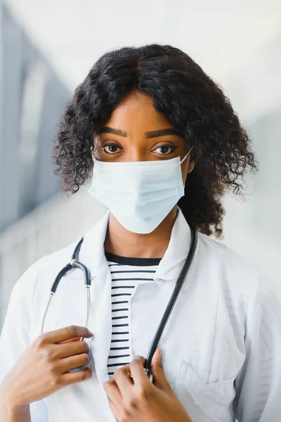 Female american african doctor, nurse woman wearing medical coat with stethoscope and mask. Happy excited for success medical worker posing on light background. Pandemia concept, covid 19