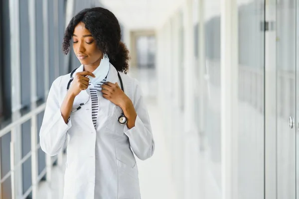 Female american african doctor, nurse woman wearing medical coat with stethoscope and mask. Happy excited for success medical worker posing on light background. Pandemia concept, covid 19