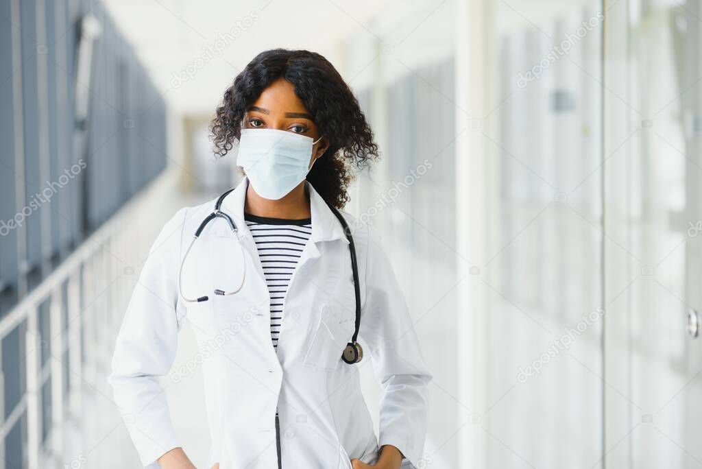 Female american african doctor, nurse woman wearing medical coat with stethoscope and mask. Happy excited for success medical worker posing on light background. Pandemia concept, covid 19